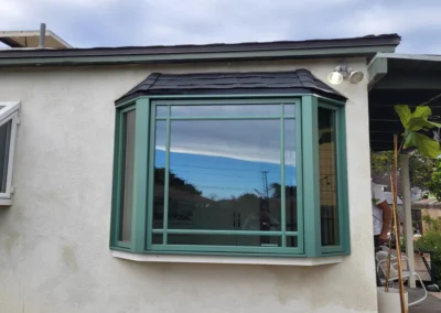 A modern bay window with green trim reflects the sky and surrounding landscape, set against a light-colored wall.